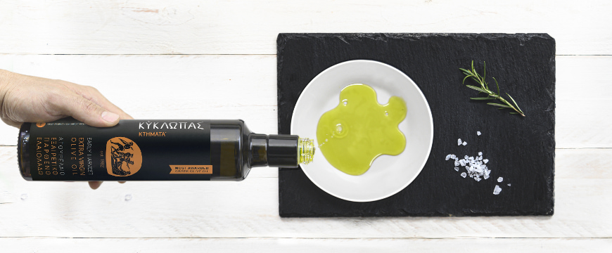 A hand pouring olive oil from a Kyklopas bottle onto a white plate