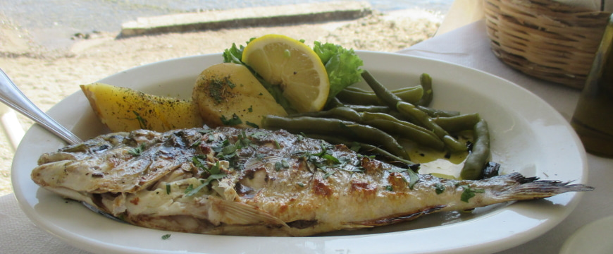 Grilled whole fish, green beans, lemon, and potato pieces with olive oil on a white oval plate