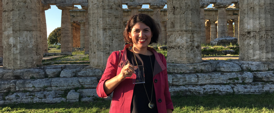 Cristina Stribacu holding her EVO IOOC award, standing in front of the base and columns of an ancient temple