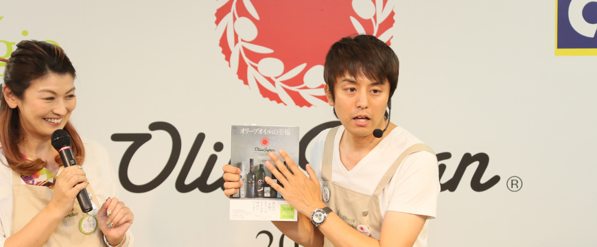 two announcers standing in front of an Olive Japan logo, one of them holding a book
