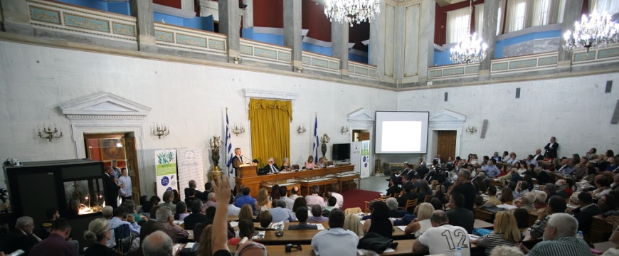 The Old Parliament auditorium in Athens, full of people, with presenters at a table and podium in front