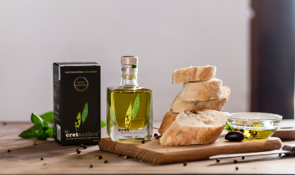 a small clear glass bottle of Cretanthos olive oil between its black box and a stack of sliced bakery bread on a cutting board
