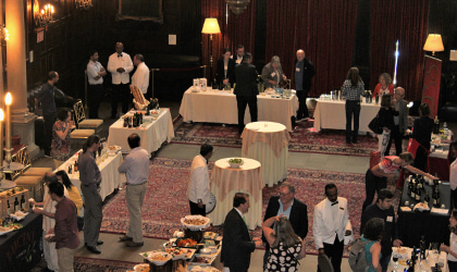 A large, high-ceilinged, wood paneled room at Harvard club with people standing around near white-clothed tables with olive oil
