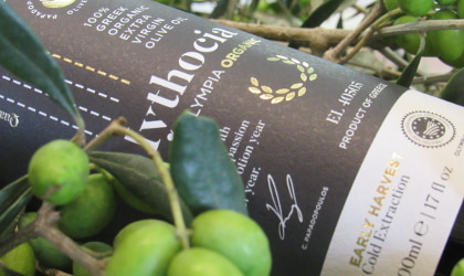 part of a bottle of the Papadopoulos Olive Oil company's Mythocia olive oil lying on its side next to green olives
