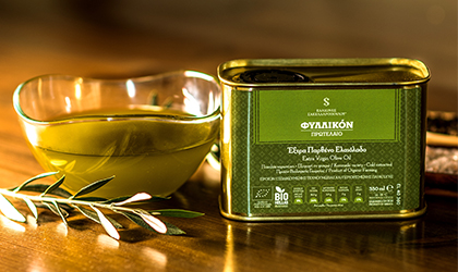 a rectangular tin of Fyllikon olive oil next to a glass bowl of olive oil, with an olive branch in front of it