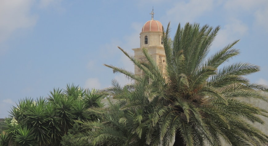 the belltower of the Toplou Monastery rishing above palm trees and yucca plants