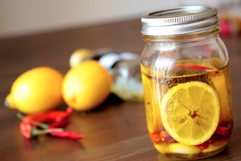 Jar containing olive oil with lemon slices and red chili peppers, with lemons and chili peppers on the table left of the jar
