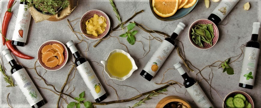 7 bottles of Pellas Nature infused olive oils lying on their sides near herbs, vegetables, and bowls of fruit, olives, and cucumber slices
