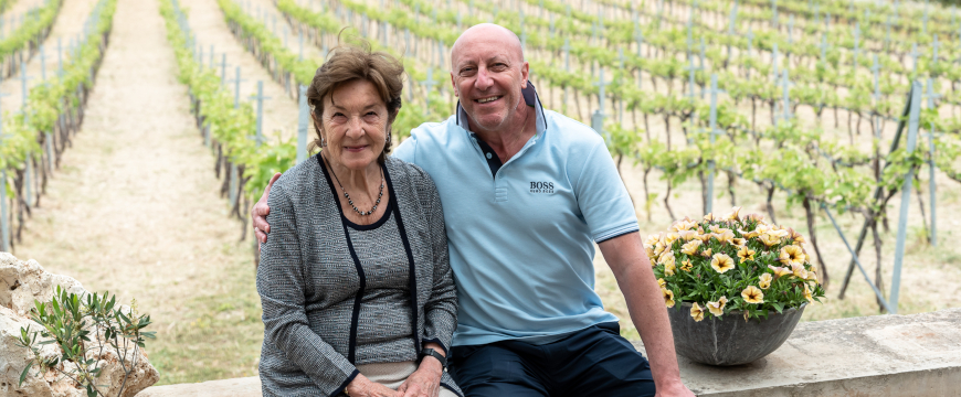 a man and a woman sitting on a wall next to some flowers, in front of a vineyard