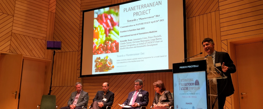 a panel of speakers on a stage, one standing at a podium, the others sitting in front of a screen that says "Planeterranean Project"