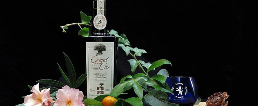 A bottle of Terra Creta Grand Cru olive oil surrounded by flowers and vines, with a blue tasting glass from the Leone d'Oro competition 