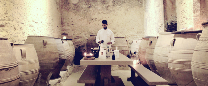 A sepia toned photo of Vasilis Leonidou behind a long table with olive oil bottles on it in a room with huge, old ceramic pots by the walls