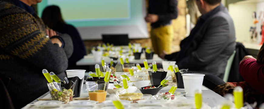 clear pipettes of olive oil stuck into various food samples on a table where people are seated for an olive oil food pairing event