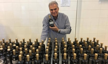 Ioannis Kampouris behind a table full of rows of his olive oil bottles, holding one of them