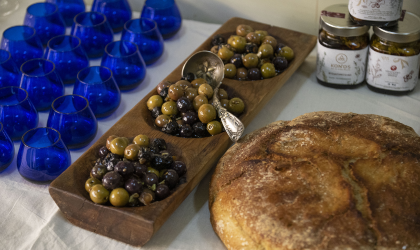 round back and green olives in a three-hole wooden dish, with blue olive oil tasting glasses on the left and a big round loaf of bread on the right, plus jars of olives behind it