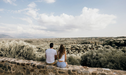 Two people sitting on a stone wall, looking at a wide expanse of olive groves below them