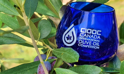 A blue olive oil tasting glass among olive branches; on the glass are the letters CIOOC and the words Canada International Olive Oil Competition in white, to the right of the competition logo