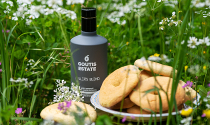 A dark gray bottle of Goutis Estate Miller's Blend olive oil near a bowl of cookies, in the grass with wildflowers