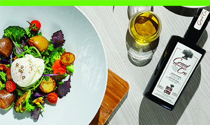 a bottle of Terra Creta Grand Cru extra virgin olive oil lying on its side next to a glass of olive oil, with a white plate full of colorful vegetables to the left