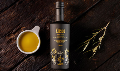 a black bottle of Krissa extra virgin olive oil lying on a wooden table with a small white bowl of golden olive oil on the left and a small olive branch on the right