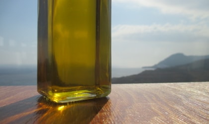 Part of a clear bottle of olive oil on a wooden table with sea and land view in the background