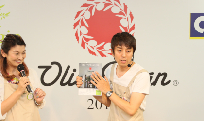A woman and a man with microphones standing in front of a large Olive Japan banner