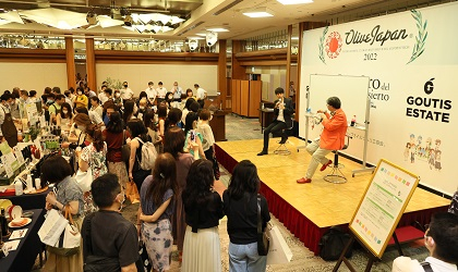 people looking at tables and at a stage with two men on it, with Olive Japan and Goutis Estate logos displayed behind the stage