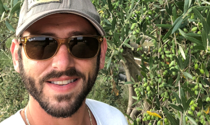 A closeup of Dino Pierrakos (face only) wearing sunglasses and a baseball cap, next to an olive tree