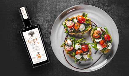 A bottle of Terra Creta Grand Cru extra virgin olive oil on the left, at an angle, and on the right a white plate with 3 pieces of bread topped with tomato, cheese, and greens, with a swirl of balsamic vinegar