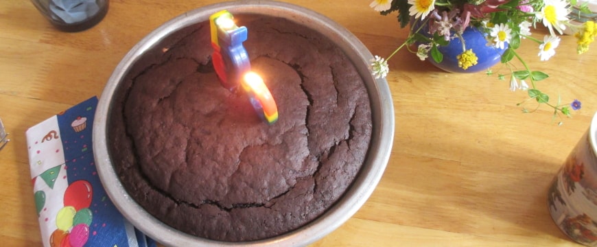 a chocolate cake in a round metal pan with two candles burning on the cake and a bouquet of flowers near it