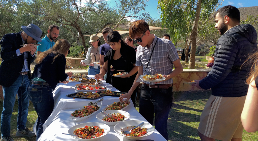 people helping themselves to food at a long table next to olive trees